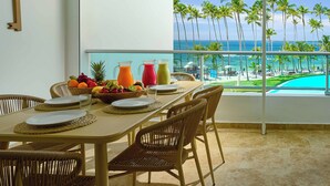 A dining area with a table set for four, a fruit bowl, and two pitchers of juice. A key is mounted on the wall. The room has a large window with a view of palm trees and a pool.