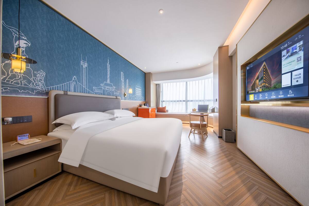 Ett modernt hotellrum med en stor säng, ett skrivbord, en stol, en tv och en väggmålning som föreställer en stadsbild.
