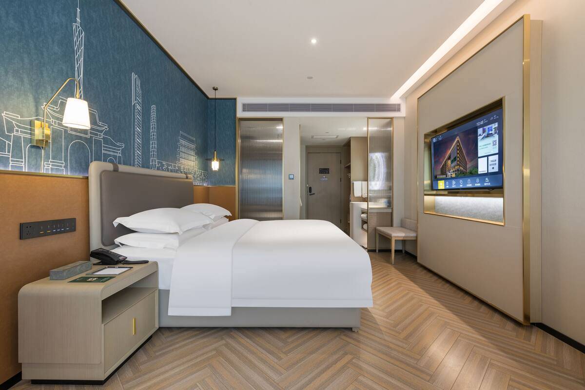 Ett modernt hotellrum med en stor säng, ett skrivbord med en lampa, en tv på väggen och en väggmålning som föreställer byggnader.