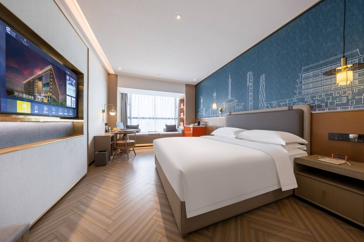 Ett modernt hotellrum med en stor säng, ett skrivbord och en tv som visar en stadsbild.
