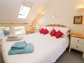 A neatly made bed with red and white pillows, a blue towel, and a wooden chair. There is a small bedside table with a lamp, a nightstand, and a framed picture on the wall. - Mill View (Cirencester)
