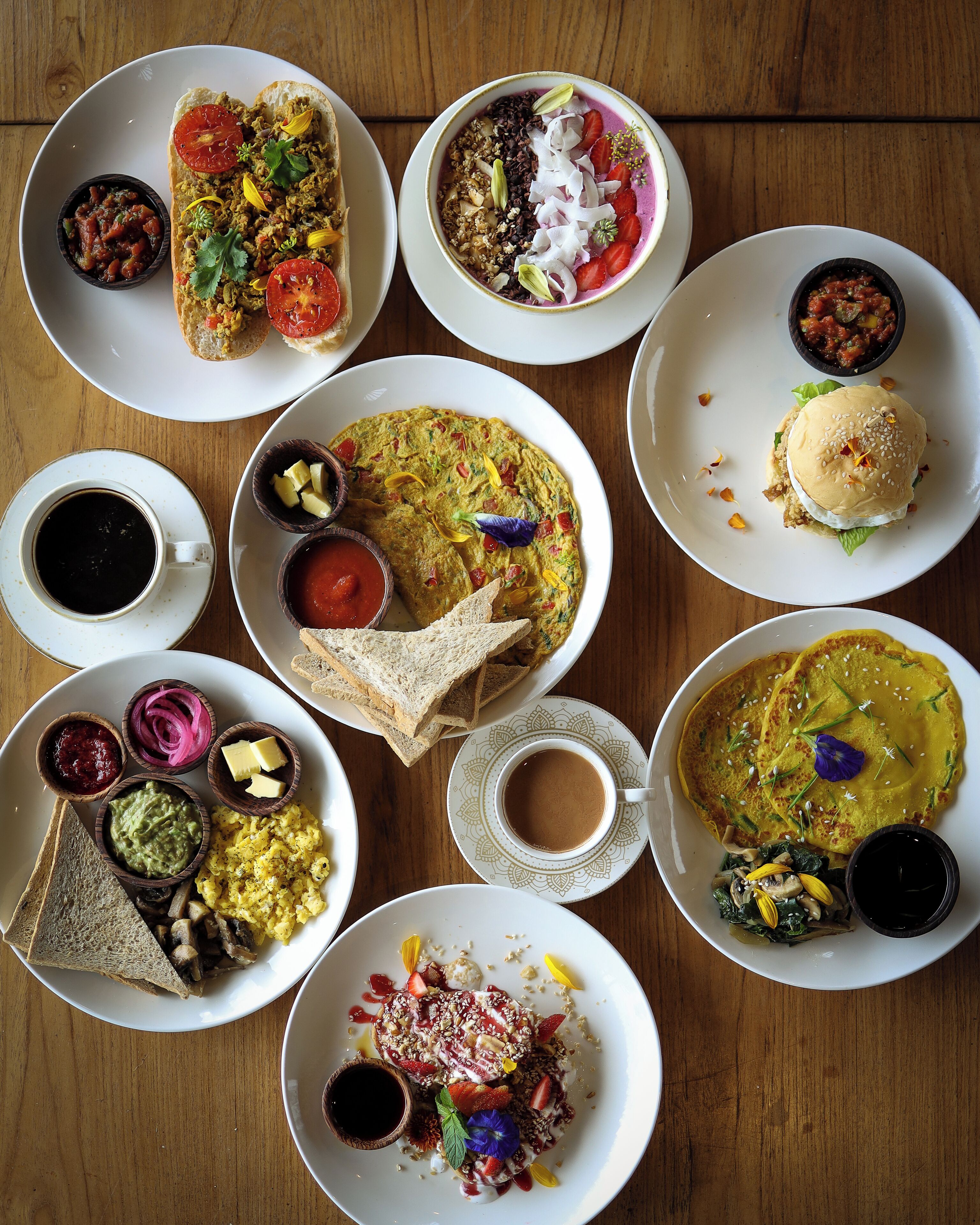A variety of dishes on a wooden table, including a sandwich, a bowl of soup, and a plate of scrambled eggs.