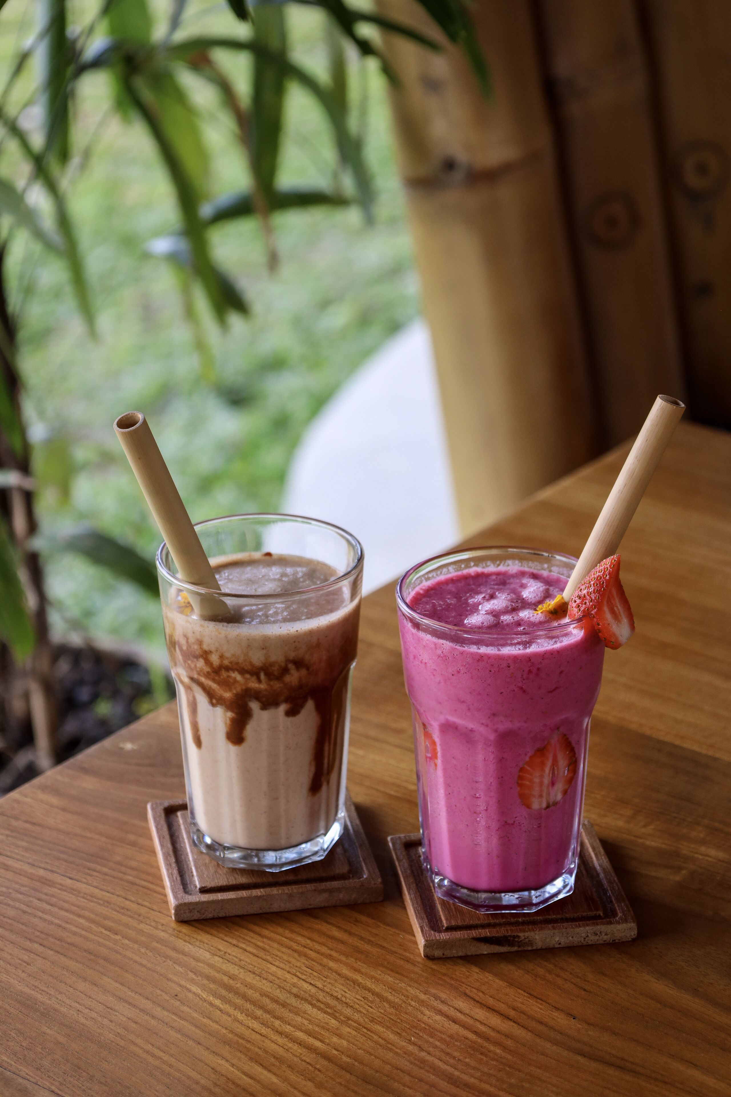 Dos vasos de batido sobre bases de madera, uno con remolino de chocolate y otro con adorno de fresa, sobre una mesa de madera con vistas a la vegetación exterior.