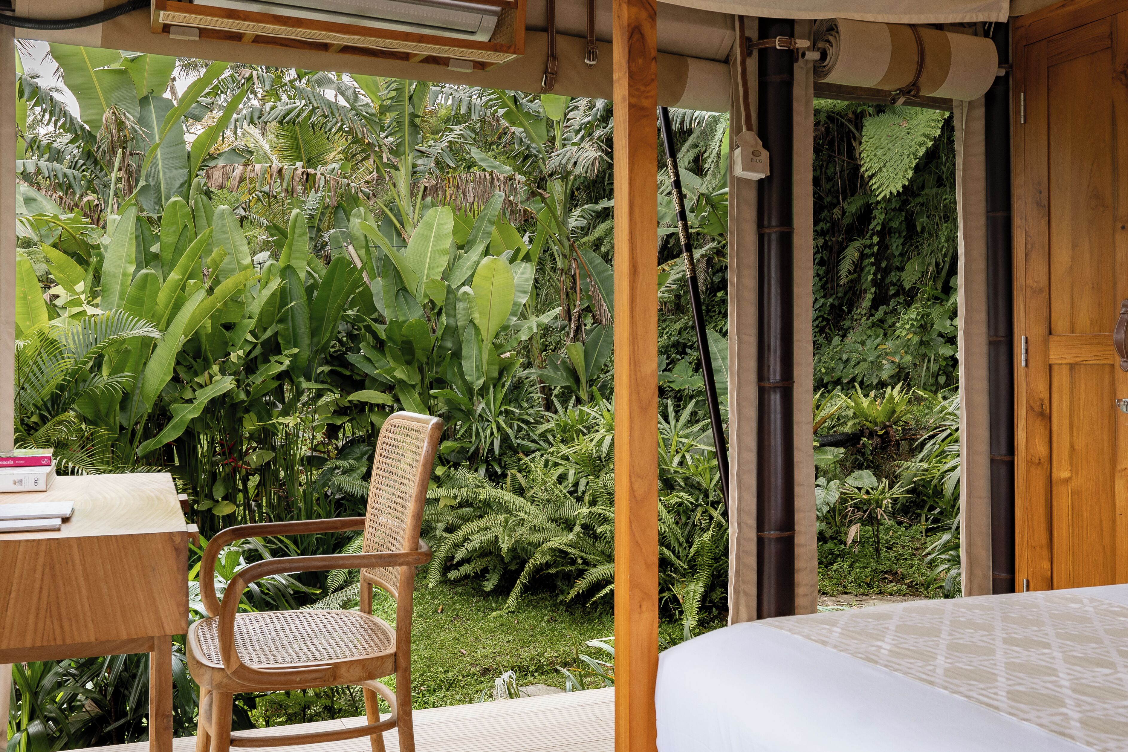 A bedroom with a bed, a wooden chair, a table with books, and a view of lush greenery outside.