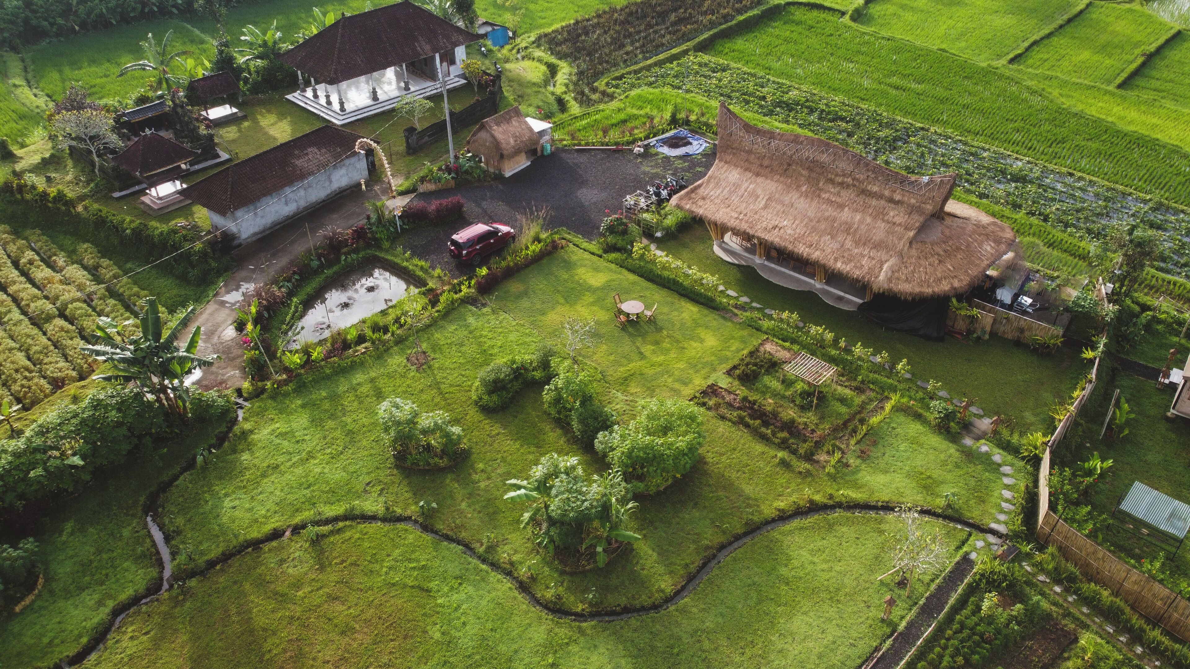 An aerial view of a traditional village with thatched-roof houses, terraced rice fields, and a central open space with a small pond and seating area.