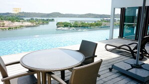 Un balcon doté d’une balustrade en verre, qui offre une vue sur une piscine et une rivière. On y trouve des chaises en osier et une table ronde sur la terrasse.