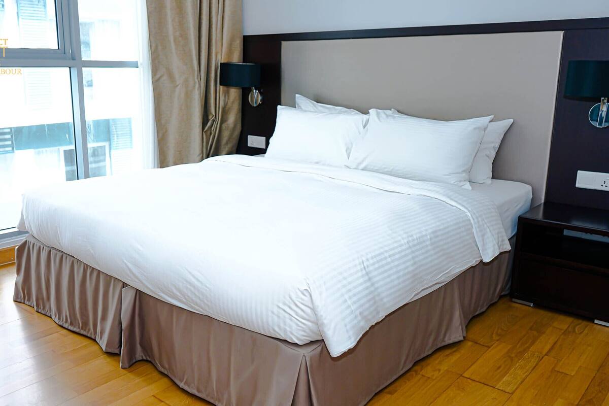 A modern hotel room with a large bed, a nightstand, a lamp, and a view of the city through large windows.