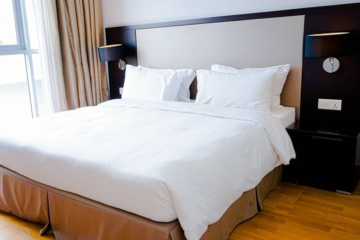 A hotel room with a large bed, a nightstand, a lamp, and a view of the city through large windows.