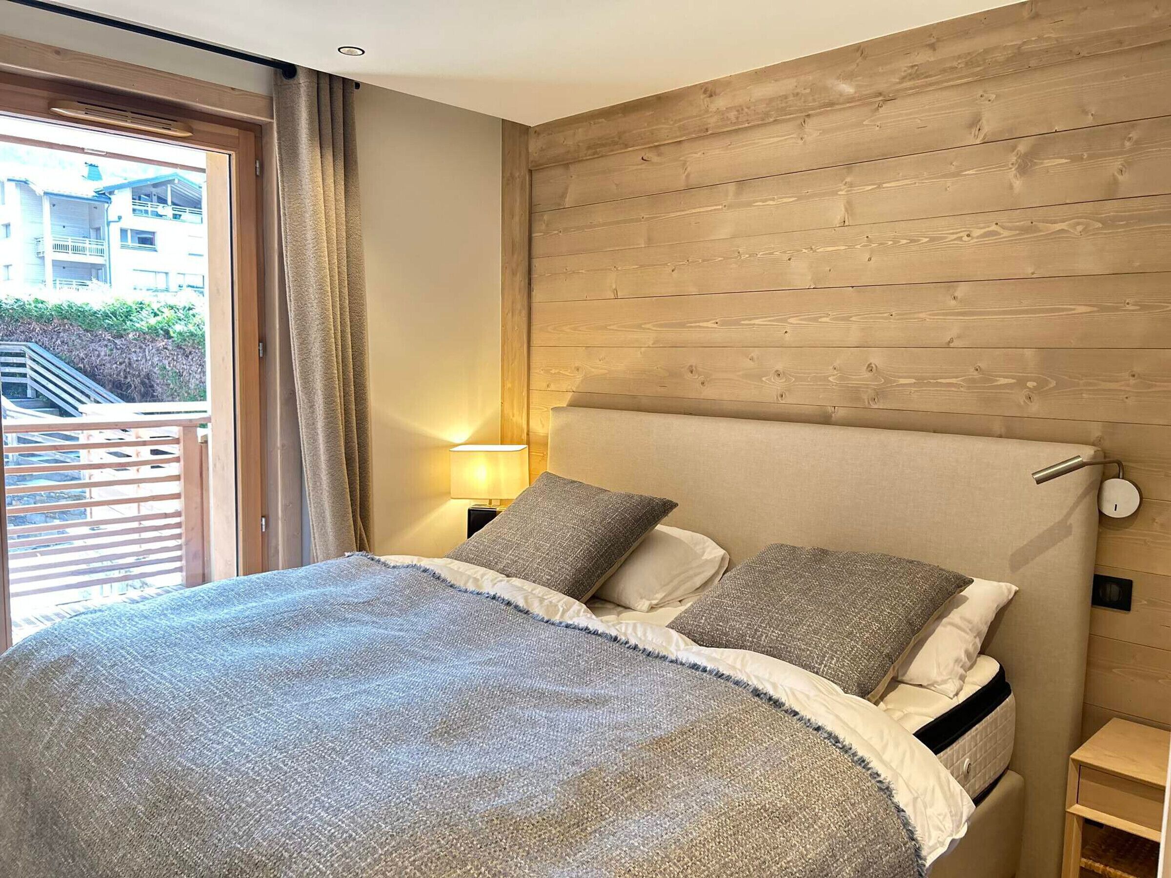 A bedroom with a wooden headboard, a large bed with a grey blanket, two pillows, a bedside table with a lamp, and a view of a balcony and buildings outside.