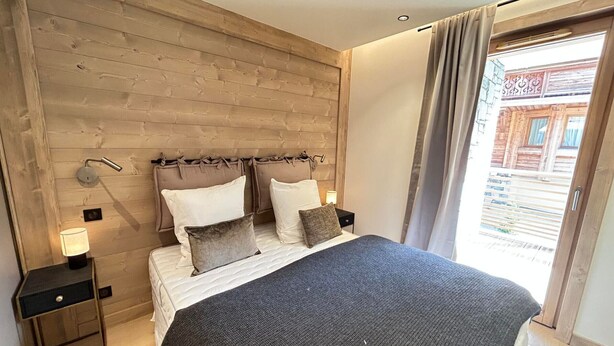 A bedroom with a wooden headboard, a bed with white and gray bedding, two bedside tables with lamps, and a view of a building through a window.