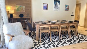 A dining area with a wooden table set for four, surrounded by chairs. There are framed pictures on the wall, a lamp, and a bookshelf with books and decorative items.