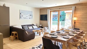 A modern living room with a sofa, a dining table set for four, and a large window with a view of a wooden cabin.