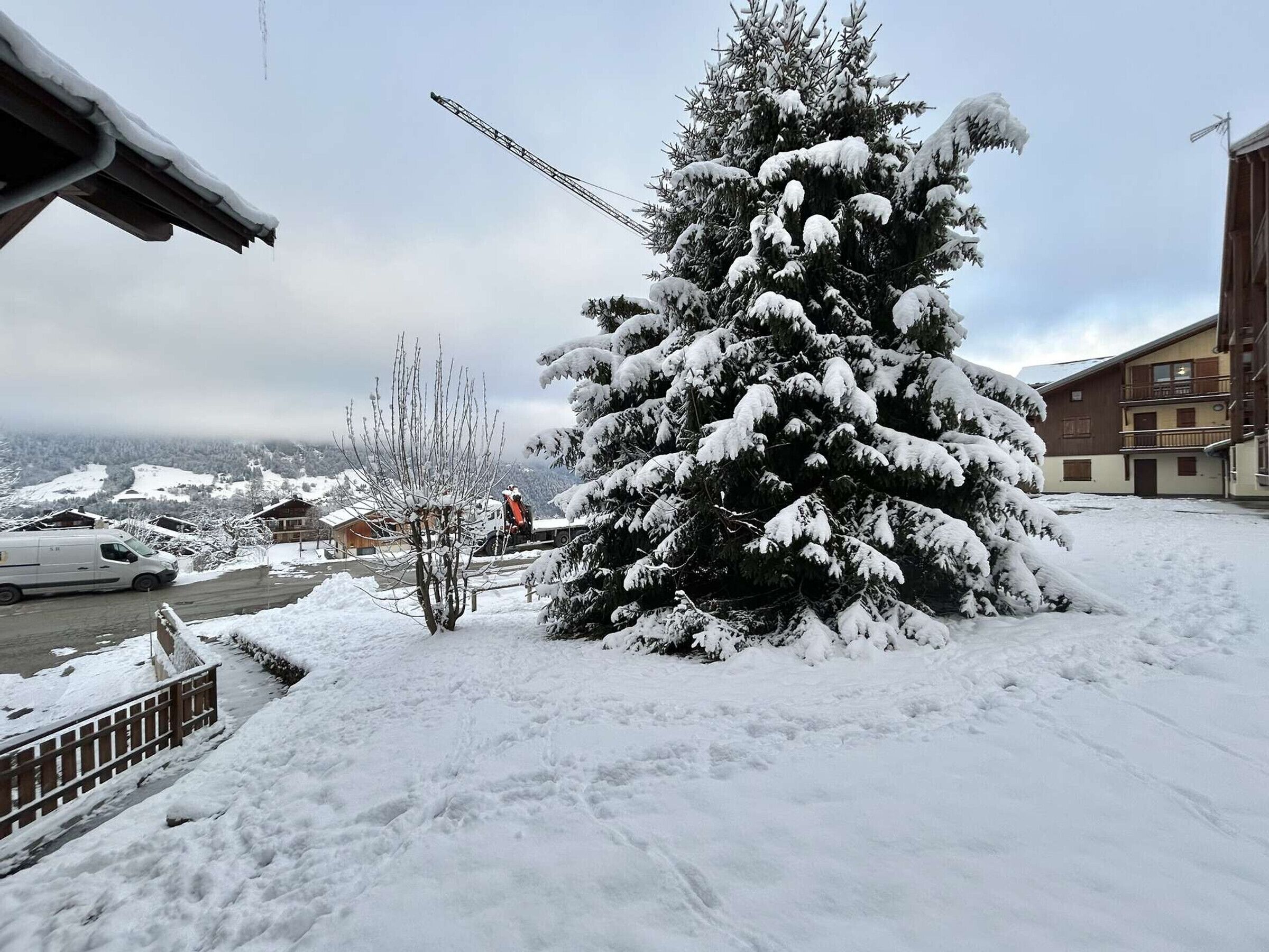 A snow-covered landscape with a large evergreen tree, a building with balconies, and a parked van.