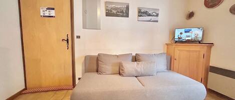 A room with a sofa, a wooden door, a television on a cabinet, and three framed pictures on the wall.
