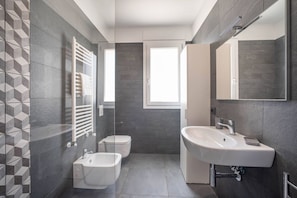 A modern bathroom with a bidet, toilet, and sink, featuring grey tiles and a window.