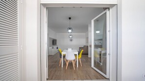 A modern interior with a dining area featuring a white table and yellow chairs, a kitchen with a glass door, and a ceiling light fixture. - WePuglia - LOI20 - Apt Maestrale (Monopoli)