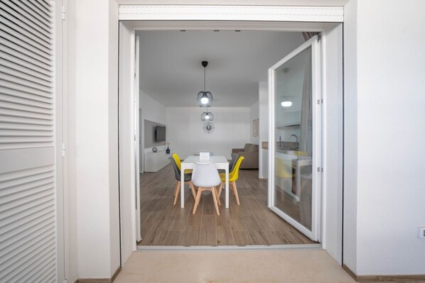 Un intérieur moderne avec un coin repas comprenant une table blanche et des chaises jaunes, une cuisine dotée d’une porte en verre et un luminaire au plafond.