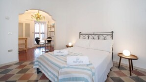 A hotel room with a bed, two bedside tables, a chandelier, a desk with a chair, and a view through an archway to another room.