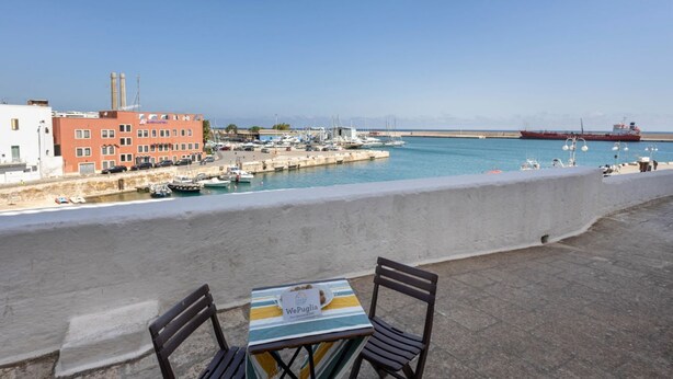 A seaside terrace with a table and chairs, overlooking a marina with boats and a large ship in the distance.