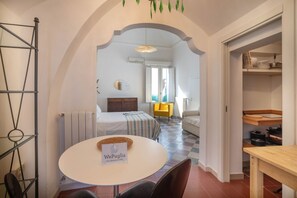 Une chambre d’hôtel compacte comprenant un lit, une table à manger, une chaise, une petite table avec un panier, et un coin cuisine avec des étagères.