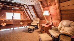 Ein gemütliches Schlafzimmer im Dachgeschoss mit schrägen Wänden, Holzwänden und einem großen Fenster. Zwei Stühle, ein kleiner Tisch und eine Lampe sind vorhanden.