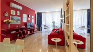 A modern living room with red walls, a red sofa, and a glass coffee table. There is a bookshelf with books and decorative items, and a flat-screen TV on a stand. - notaMI - Twins Apartment  (Milan)