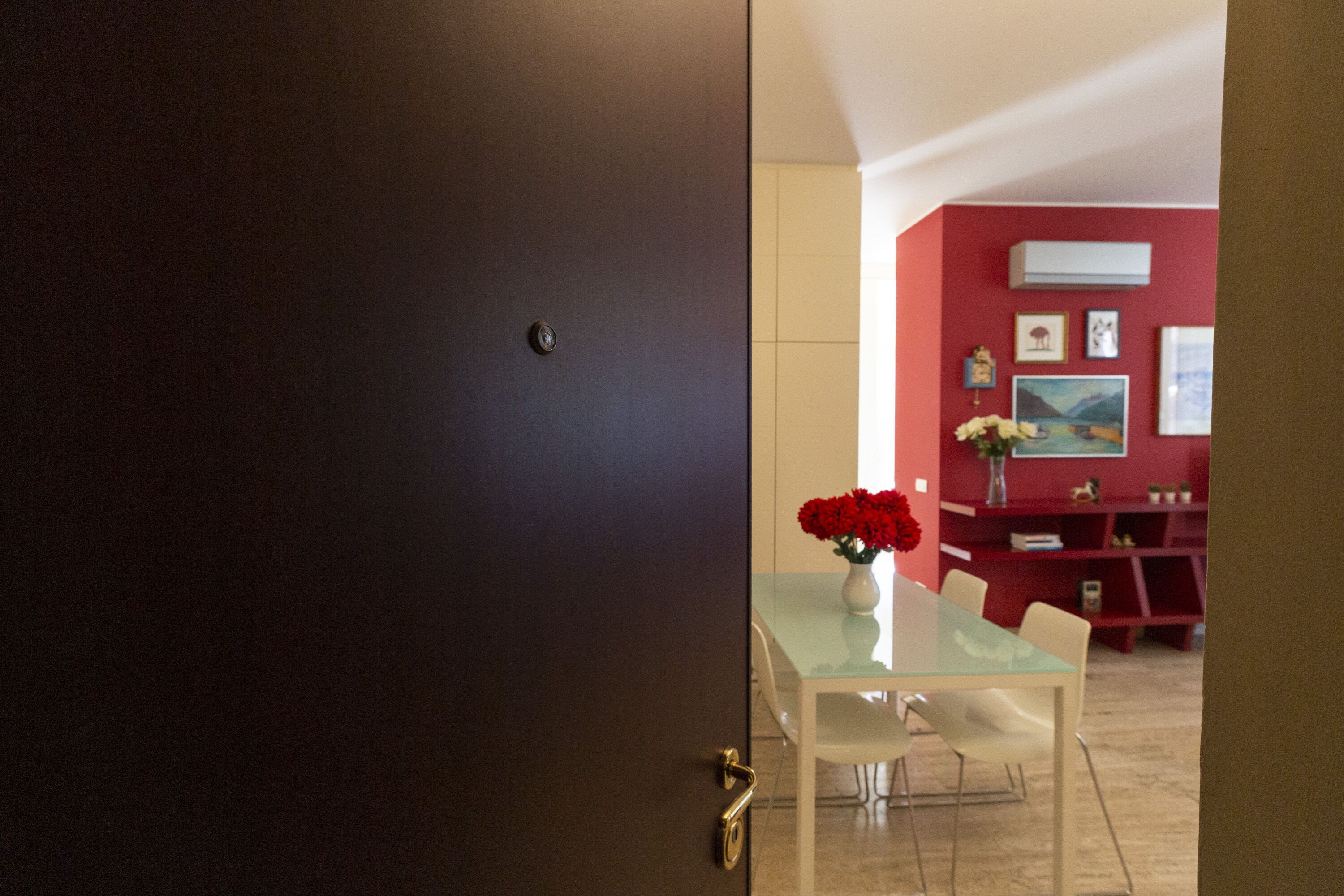 A modern interior with a dark door, a glass table with chairs, and a red wall with artwork.