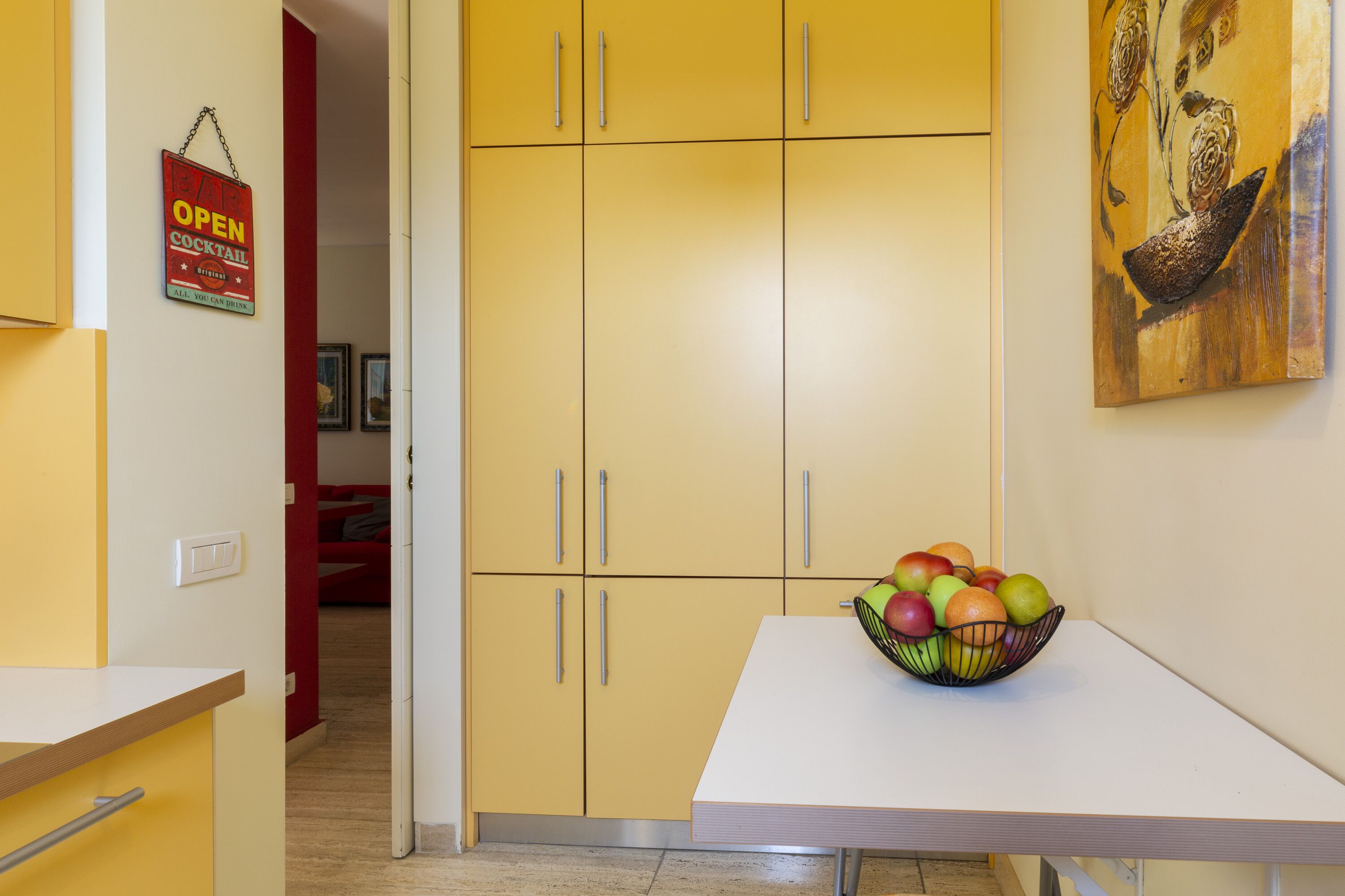 Une pièce avec des meubles de rangement jaunes, une table blanche sur laquelle se trouve un panier de fruits, et un tableau accroché au mur.