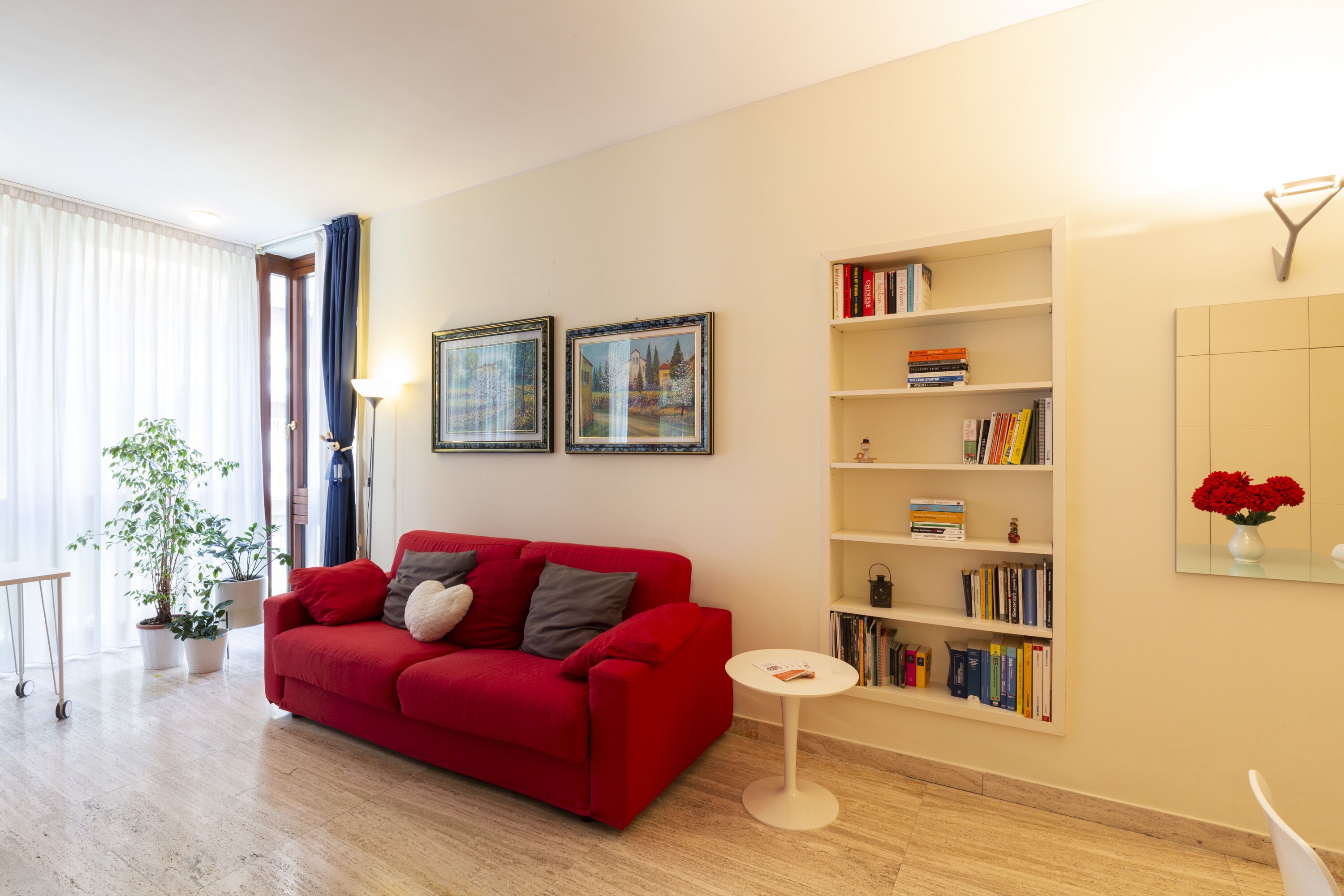 A living room with a red sofa, a white side table, and a bookshelf filled with books.