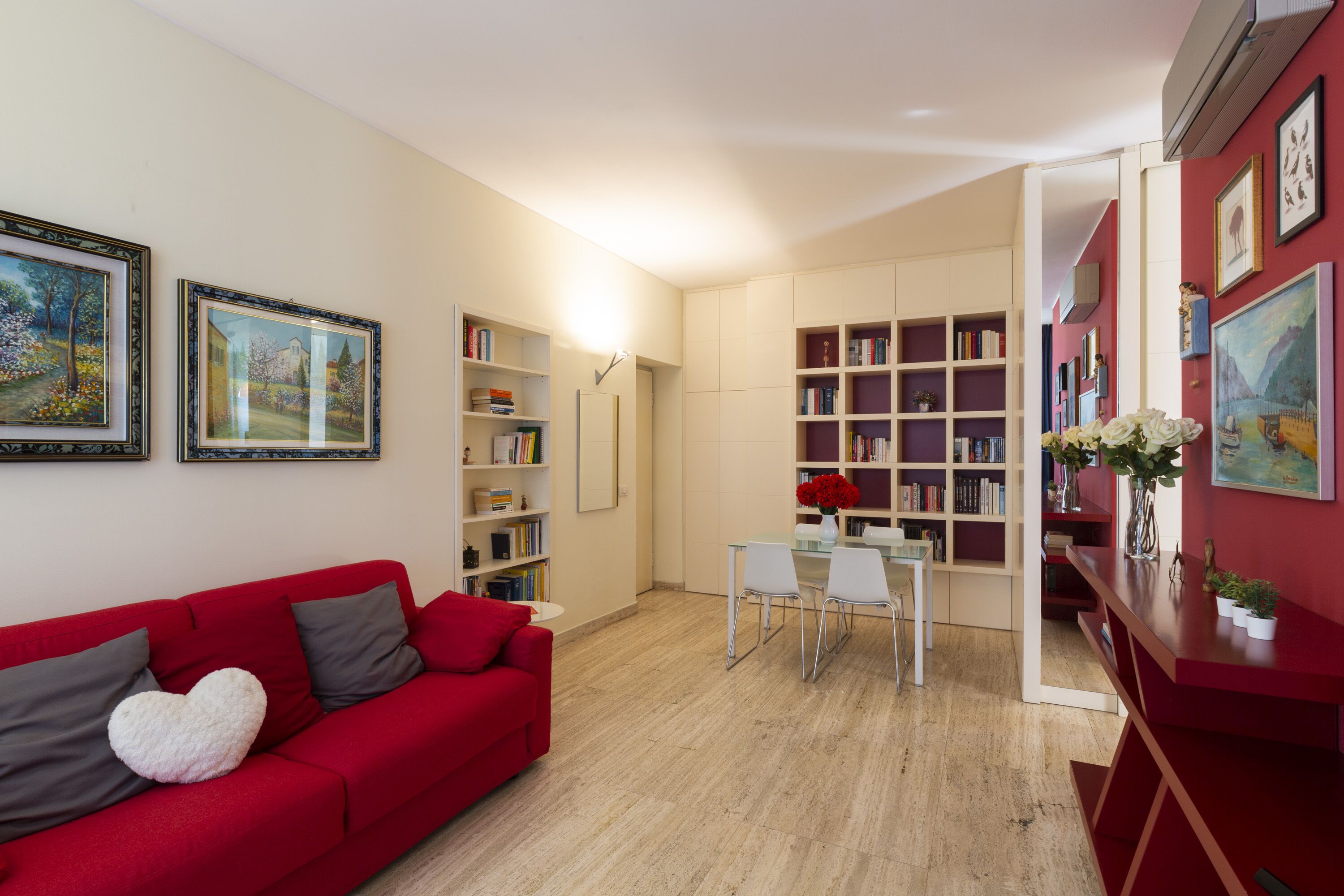 Ein Wohnzimmer mit einem roten Sofa, einem Esstisch mit Stühlen und Bücherregalen voller Bücher.