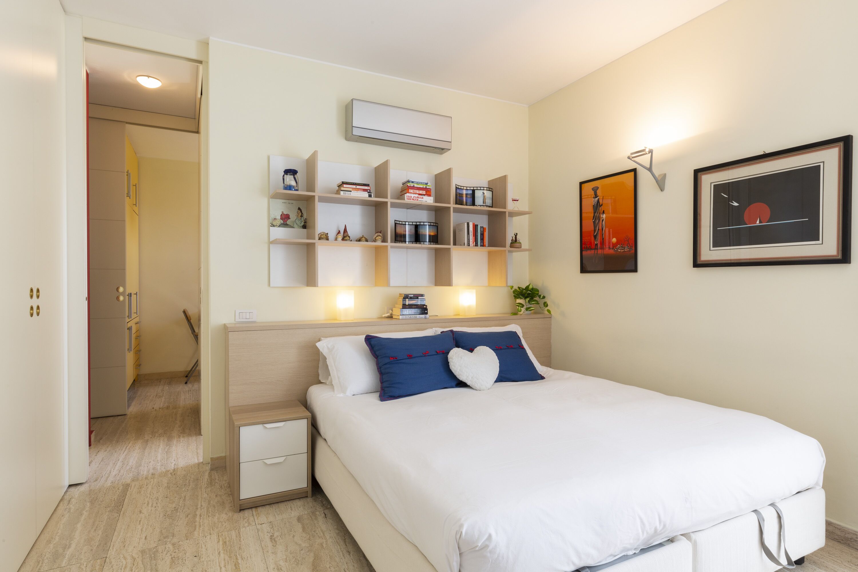 A bedroom with a bed, bedside tables, a shelf with books and decorative items, a wall-mounted air conditioner, and a framed picture.