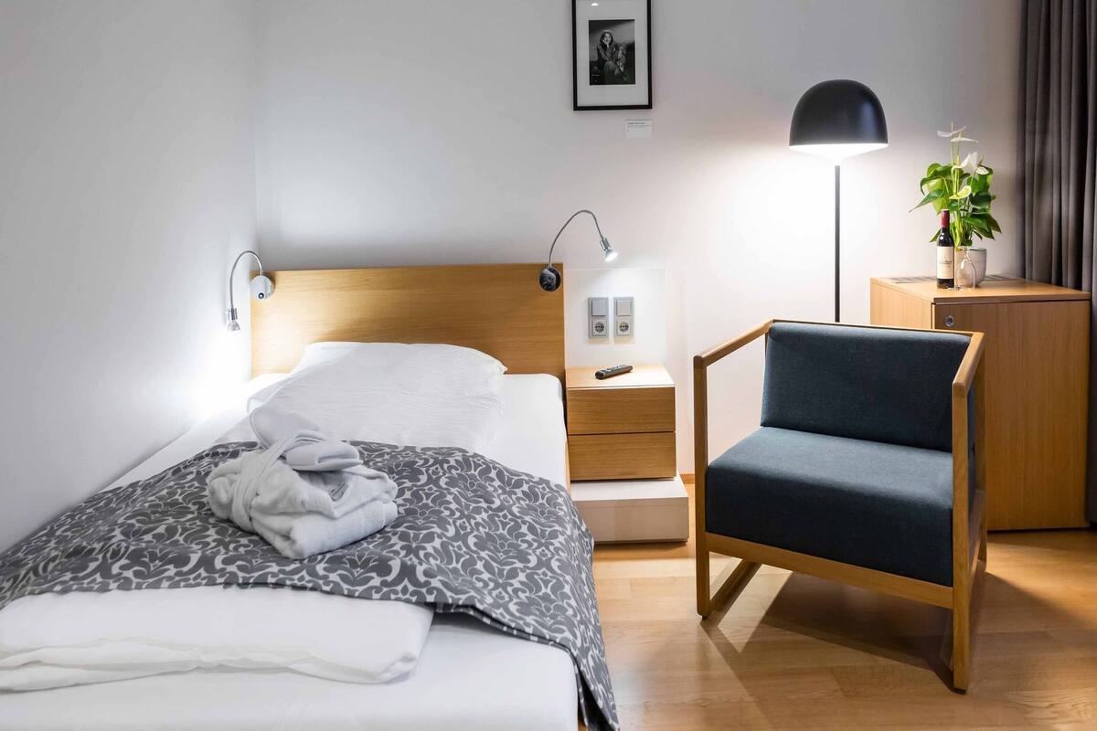 A neatly made bed with a patterned blanket, a bedside lamp, a small wooden nightstand with a remote control, a blue upholstered chair, and a potted plant on a wooden cabinet.