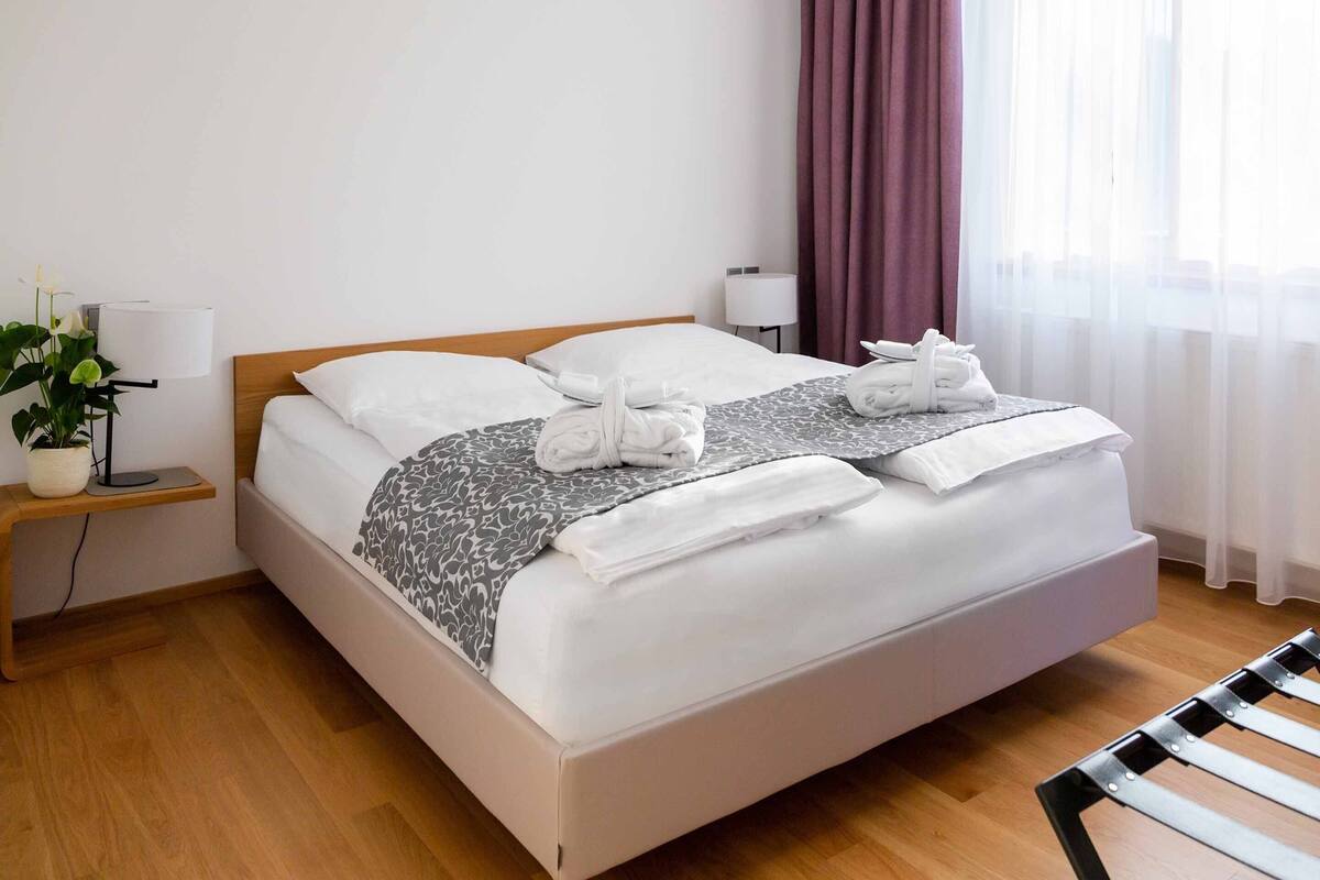 A neatly made bed with white linens and a grey patterned throw, a bedside lamp, a small table with a plant, and a chair.