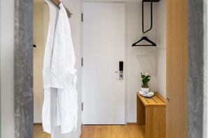 A hotel room with a wooden floor, a white door, a robe hanging on a rack, a small wooden table with a potted plant, and a black hanger on the wall.