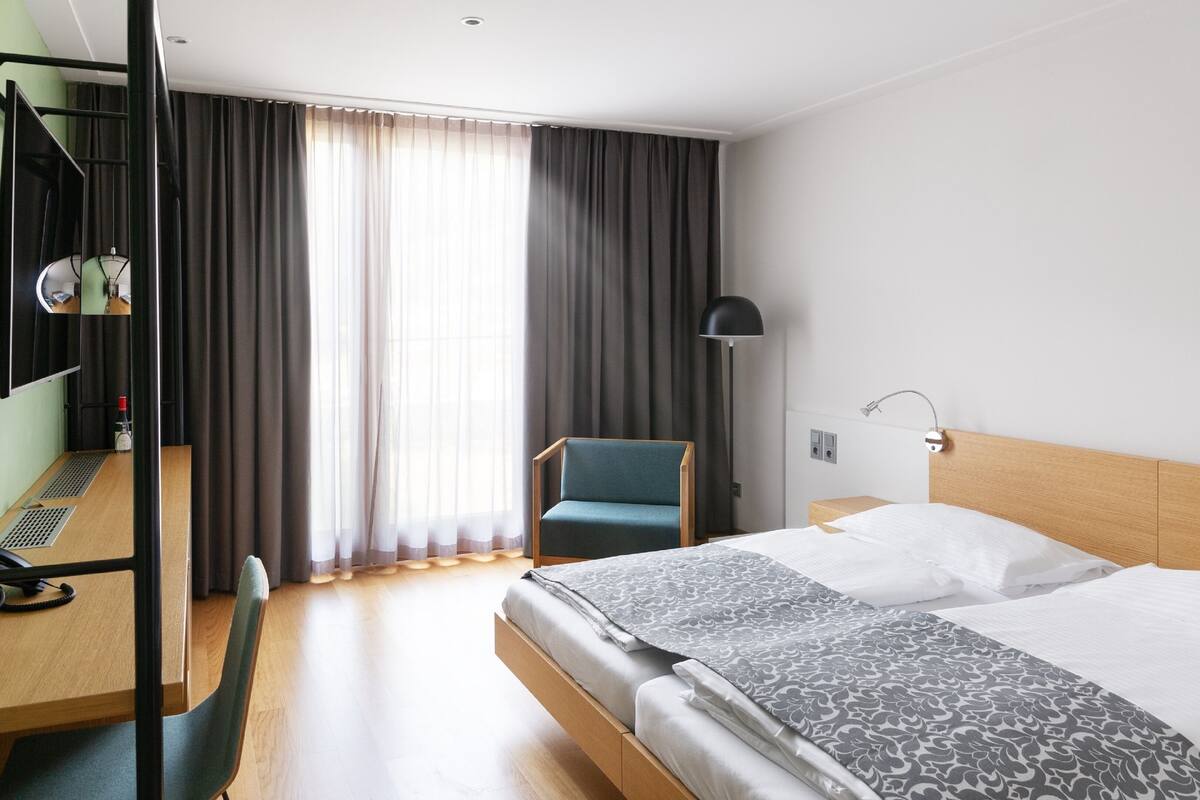 A hotel room with a bed, a desk, a chair, a lamp, and a window with curtains.