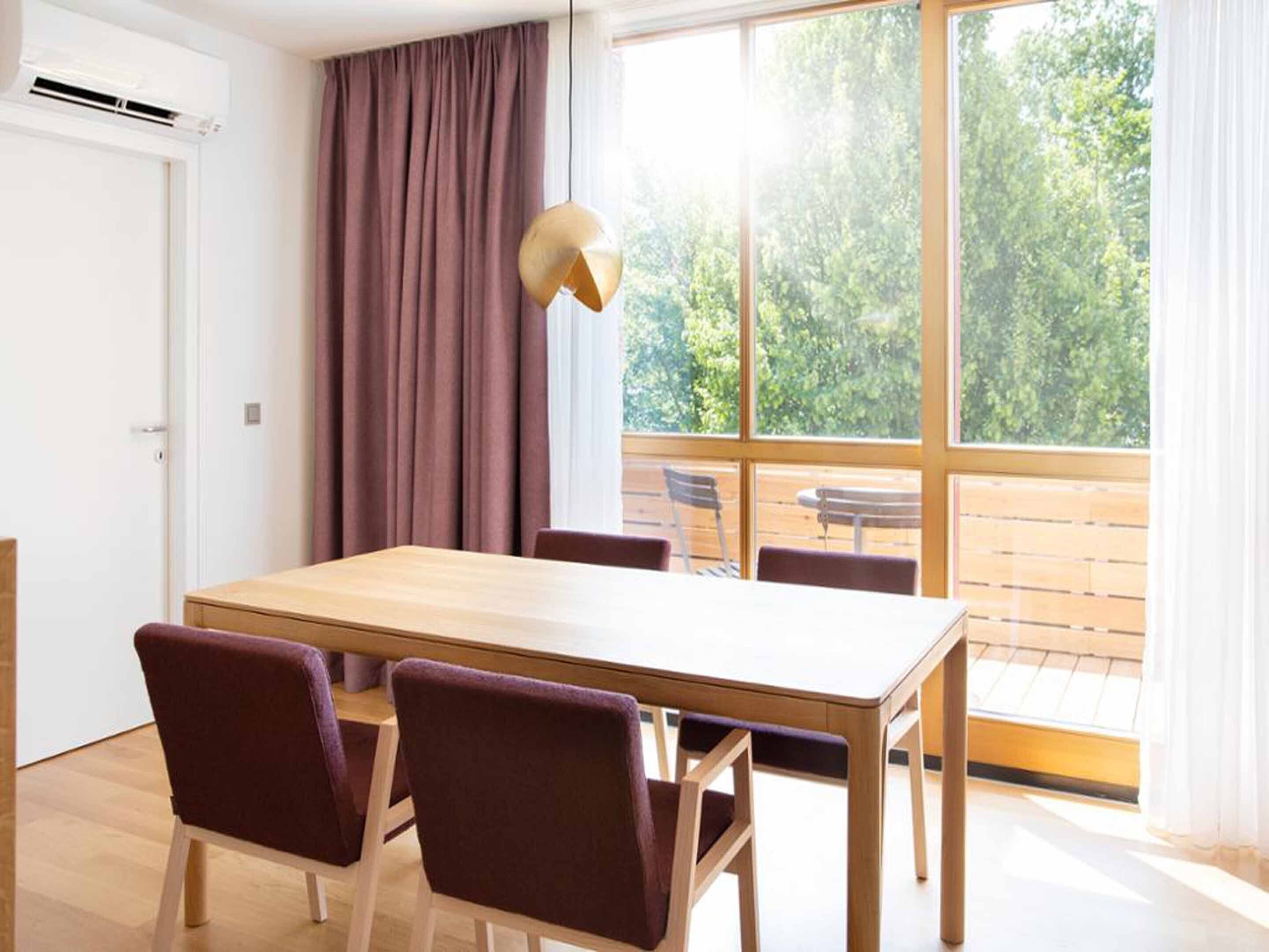 A dining area with a wooden table and four chairs, a hanging light fixture, and a view of a balcony with outdoor furniture.
