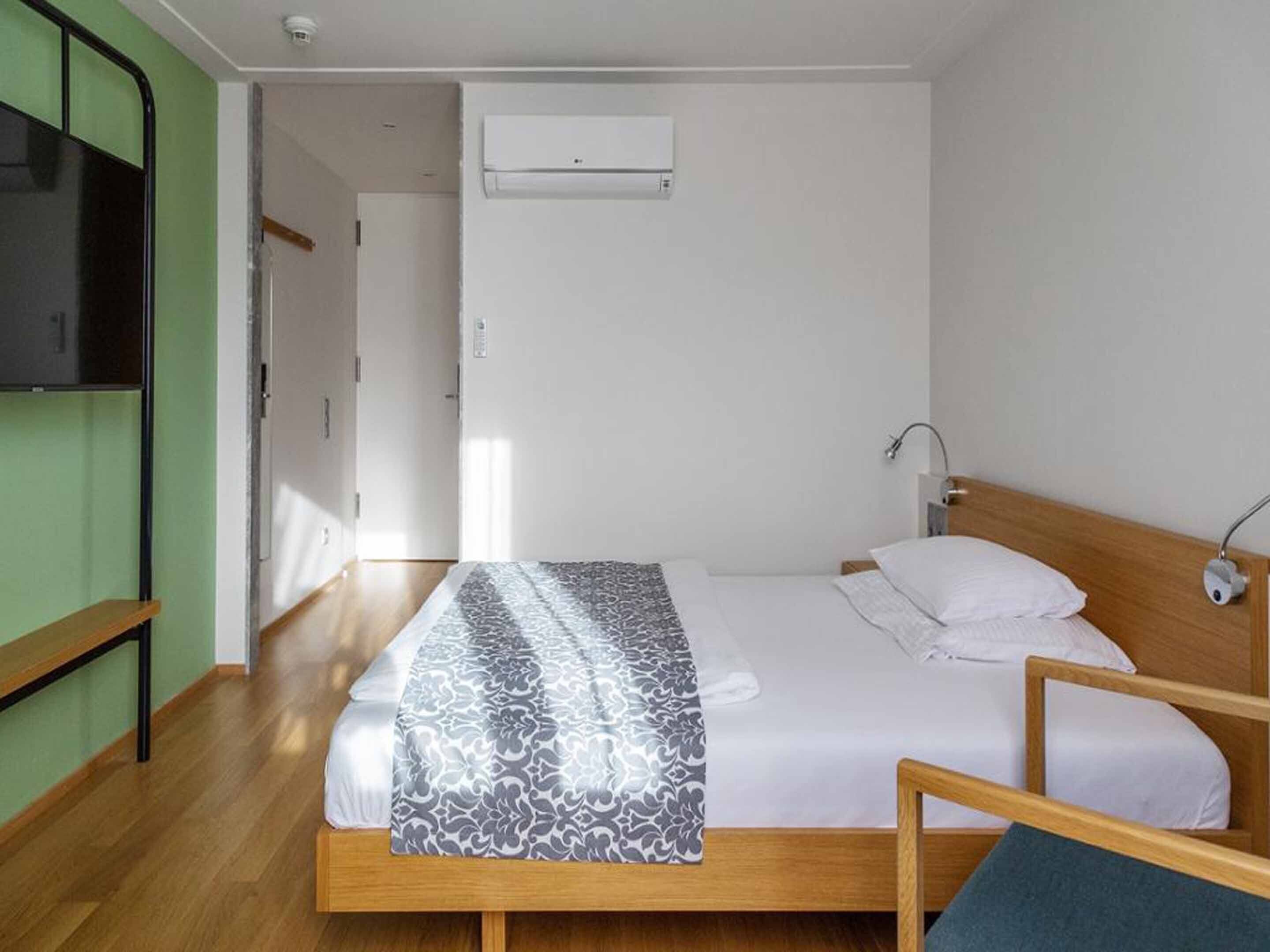 A single-bed room with a wooden frame, a patterned bedspread, a bedside table with a lamp, and a wall-mounted air conditioner.