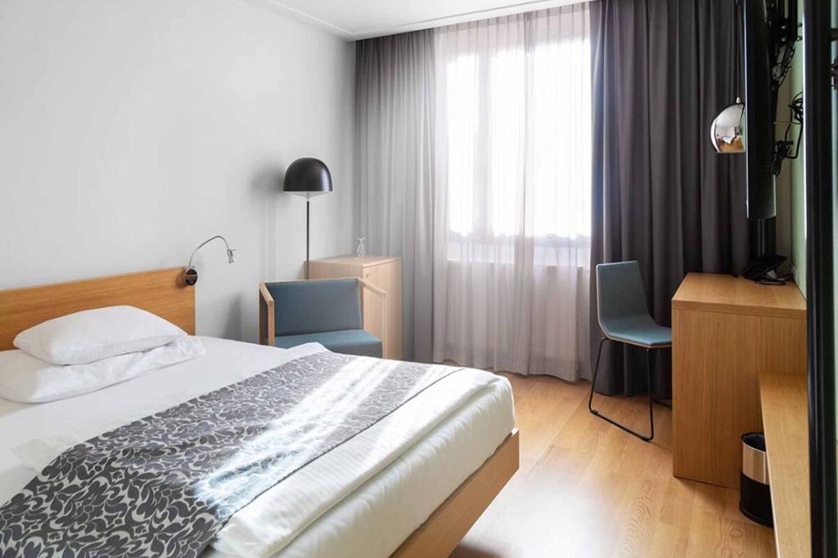 A hotel room with a bed, a desk, a chair, a lamp, and a view of the outdoors through the window.