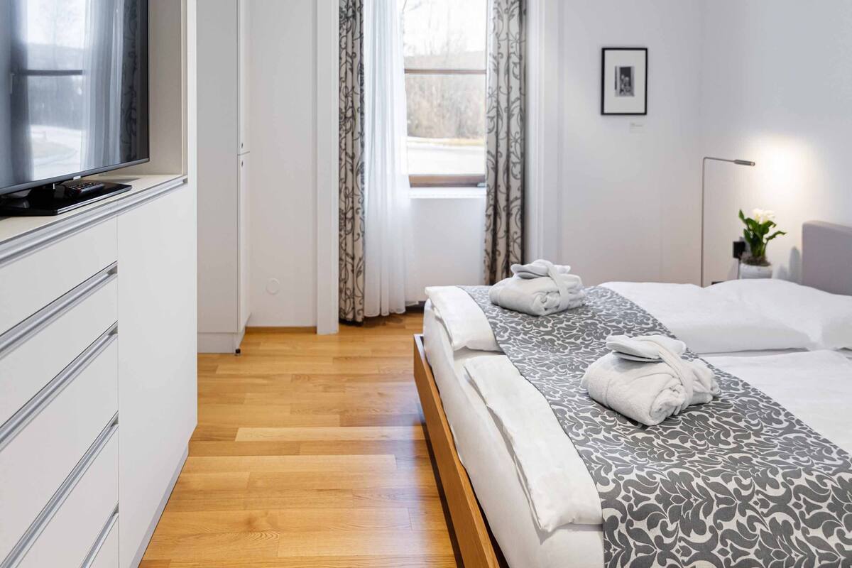 A modern hotel room with a wooden floor, a bed with patterned bedding, a TV on a cabinet, and a window with curtains.