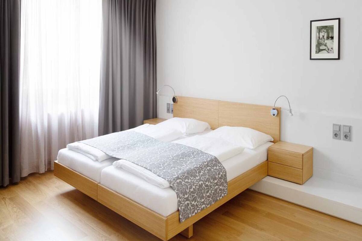 A modern bedroom with a wooden bed, white linens, a grey patterned throw, a bedside table with a lamp, and a framed picture on the wall.