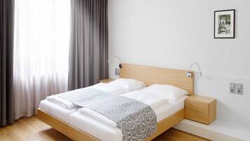A modern bedroom with a wooden bed, white linens, a grey patterned throw, a bedside table with a lamp, and a framed picture on the wall.