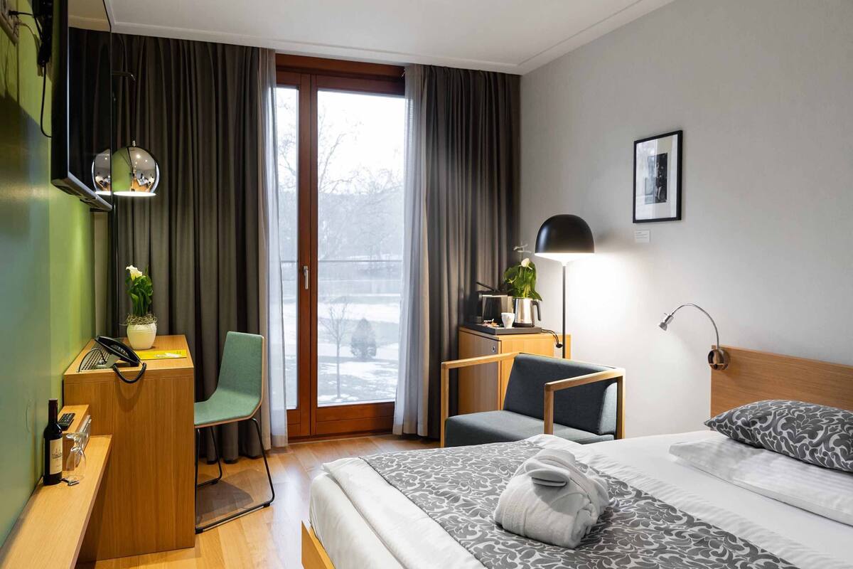 A hotel room with a bed, a desk with a telephone, a chair, a desk lamp, and a window with curtains.