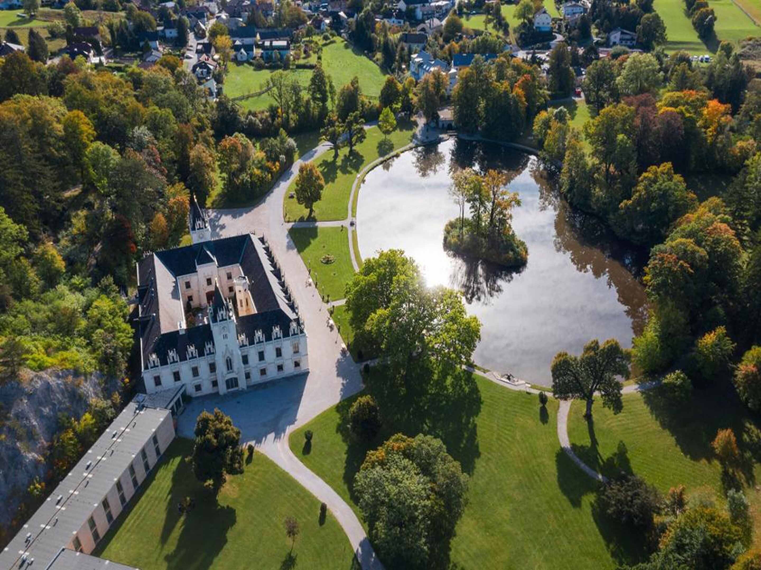 Eine Vogelperspektive auf ein historisches Gebäude mit einer symmetrischen Fassade, umgeben von einem gepflegten Park mit einem See und Wegen.
