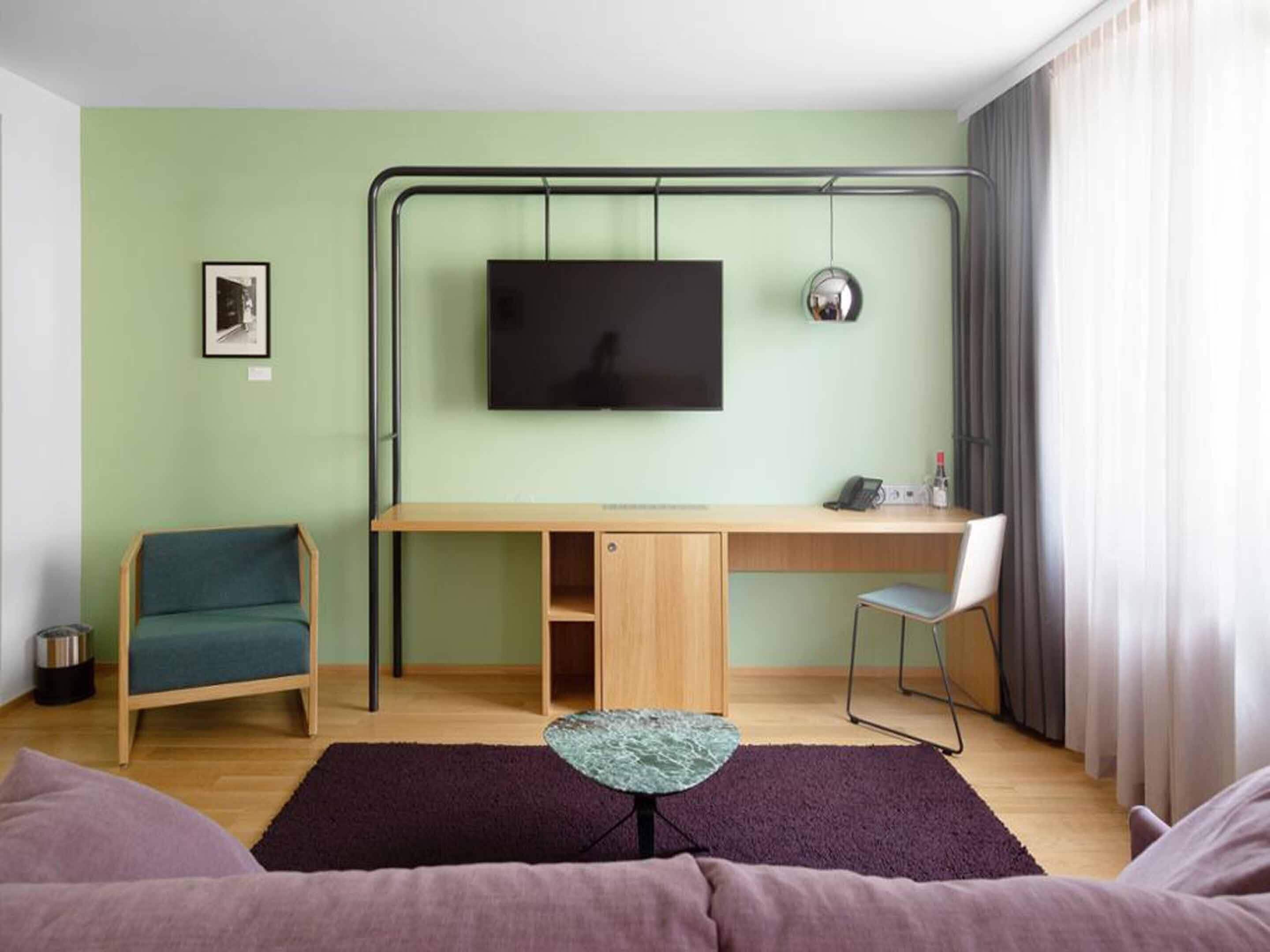 Een moderne kamer met een aan de muur bevestigde televisie, een bureau met stoel, een klein tafeltje en een bank.