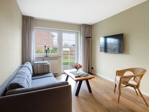 A living room with a sofa, a table with a vase of flowers, a wicker chair, and a television on the wall.