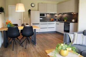Uma cozinha moderna com armários brancos, micro-ondas e fogão. Há uma área de refeições com uma mesa de madeira e cadeiras pretas.