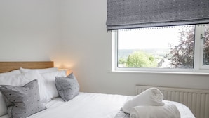 Een net opgemaakt bed met wit en grijs beddengoed, een houten hoofdbord en een raam met uitzicht op groen en gebouwen.
