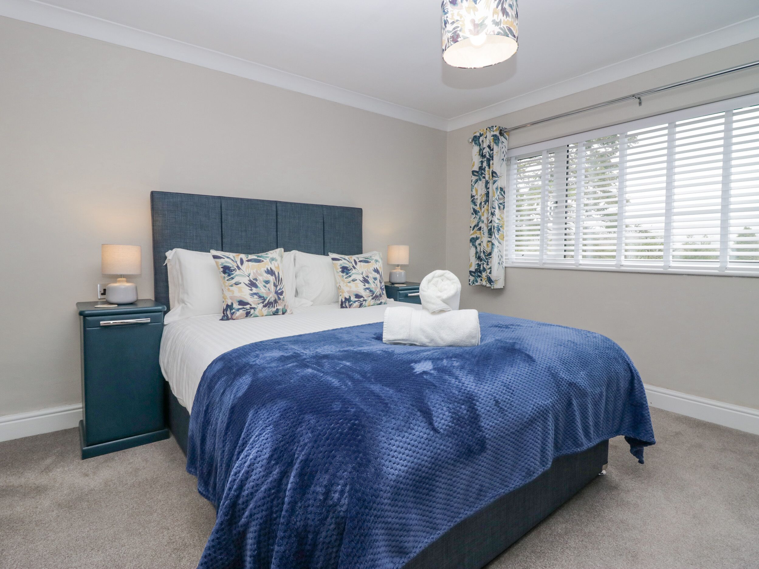 Un dormitorio con cama, mesitas de noche, lámpara, ventana con persianas y almohada decorativa.