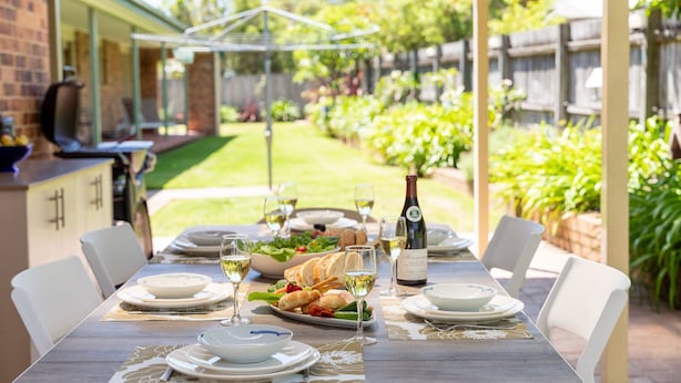 Un área de comedor al aire libre con una mesa dispuesta para cuatro personas, rodeada de sillas. Se observa una parrilla y una mesa de comedor con platos, vasos y una botella de vino.
