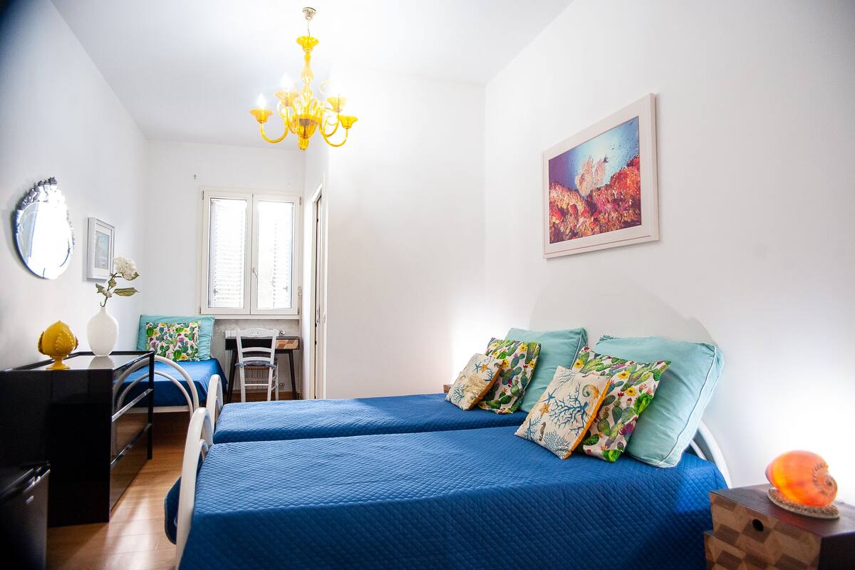A bedroom with two beds, a desk, a chair, a mirror, a chandelier, and a painting on the wall.